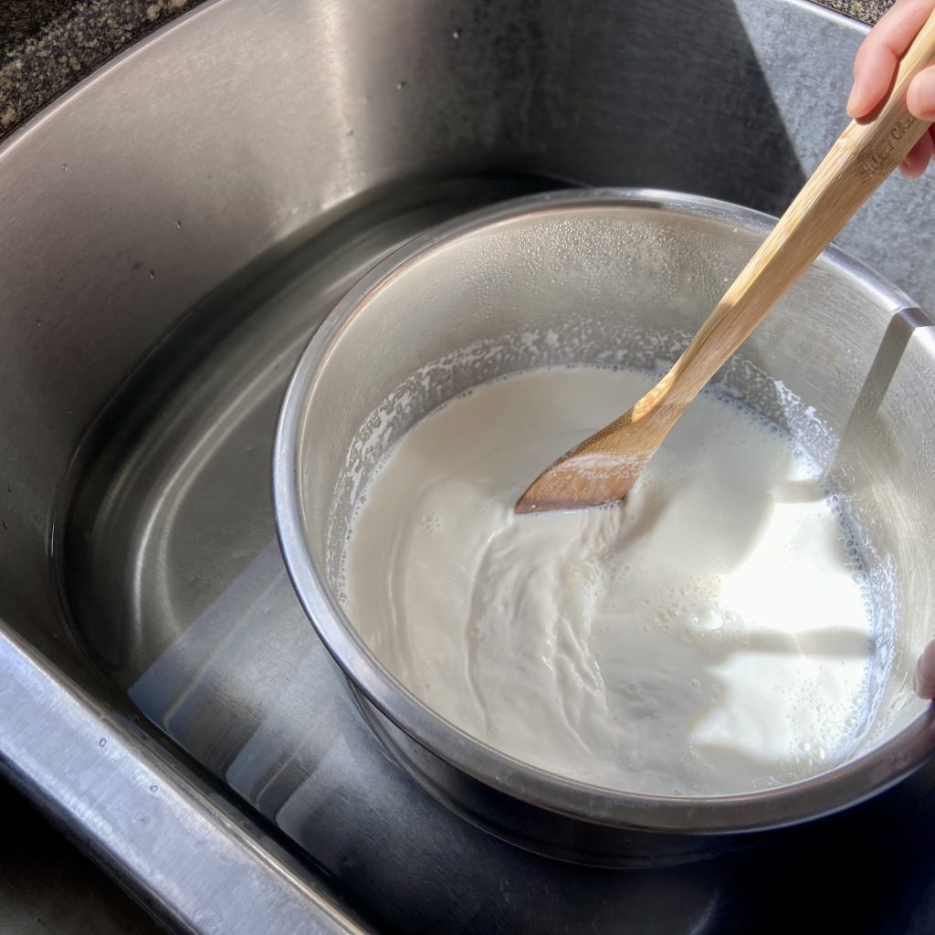 Pot of hot milk resting in a sink of cold water to cool it down while someone stirs it