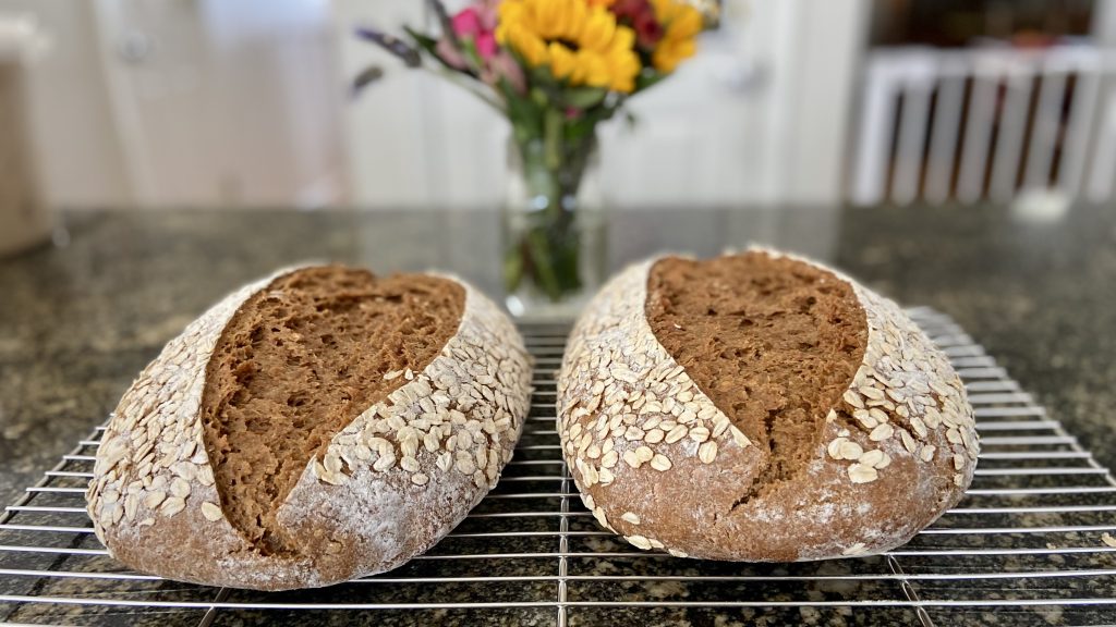 Two loaves of oatmeal rye sourdough bread resting on a cooling rack with flowers in the background