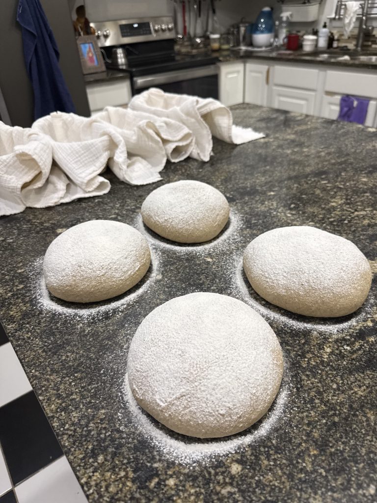 There are four loaves of bread shaped and dusted with flour, resting on a counter waiting to be placed in proofing baskets.